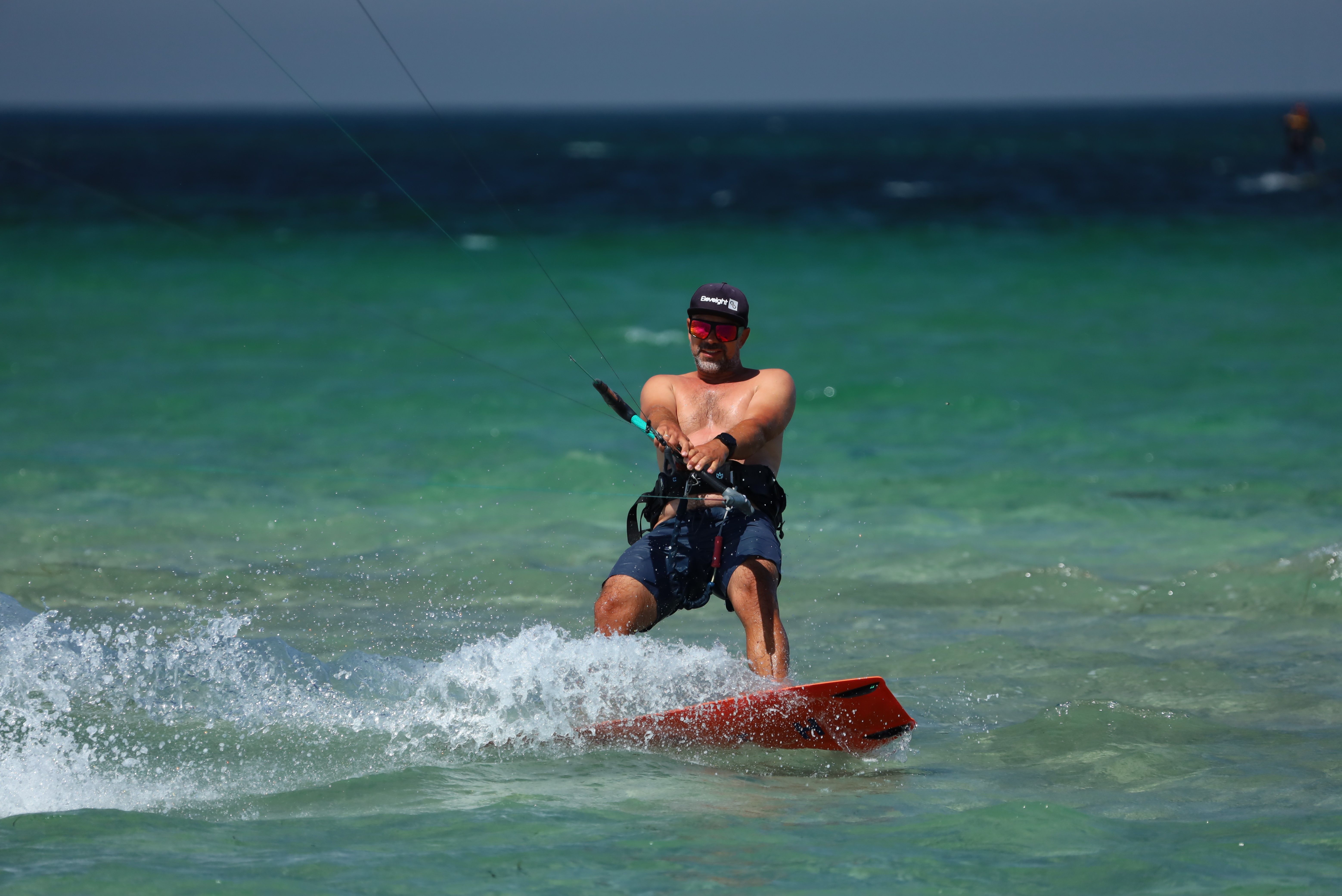 Shirtless man enjoying kiteboarding on vibrant turquoise waters, wearing cap and sunglasses.