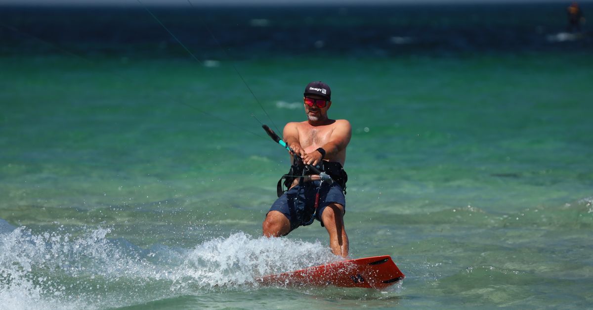 Shirtless man enjoying kiteboarding on vibrant turquoise waters, wearing cap and sunglasses.