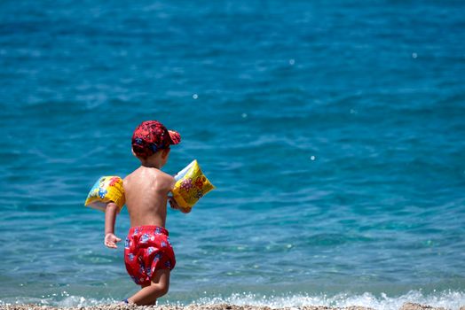 A child in swim gear plays on a sunny Greek beach, enjoying the clear blue sea.