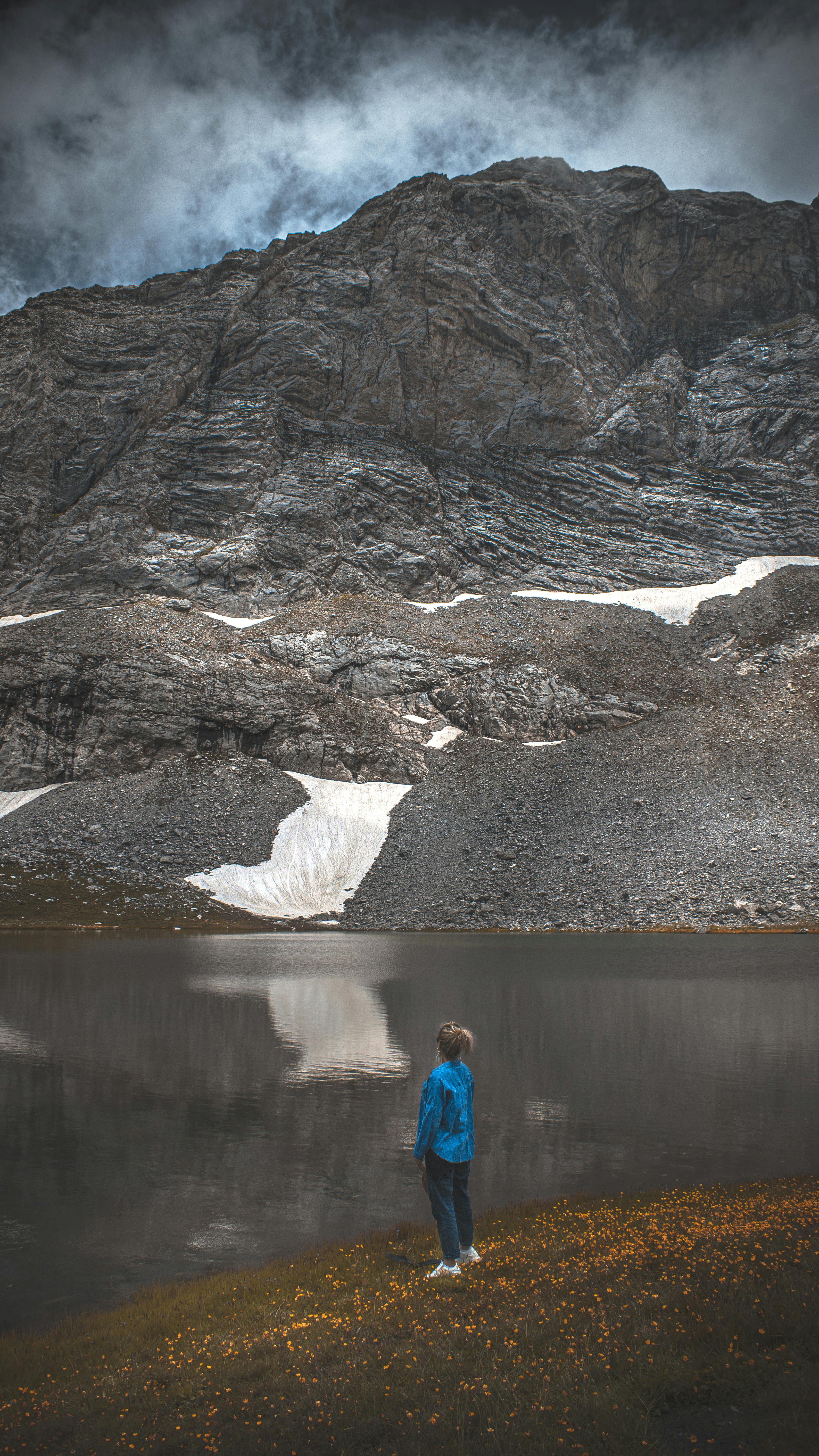 Prescription Goggle Inserts - A woman in a blue jacket stands by a tranquil mountain lake, taking in the rugged landscape.