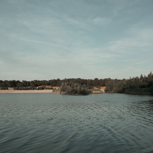 Free Tranquil lake scene with surrounding lush trees under a blue sky, perfect for nature lovers. Stock Photo
