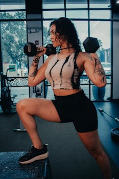 A tattooed woman exercising with dumbbells in a modern gym setting.