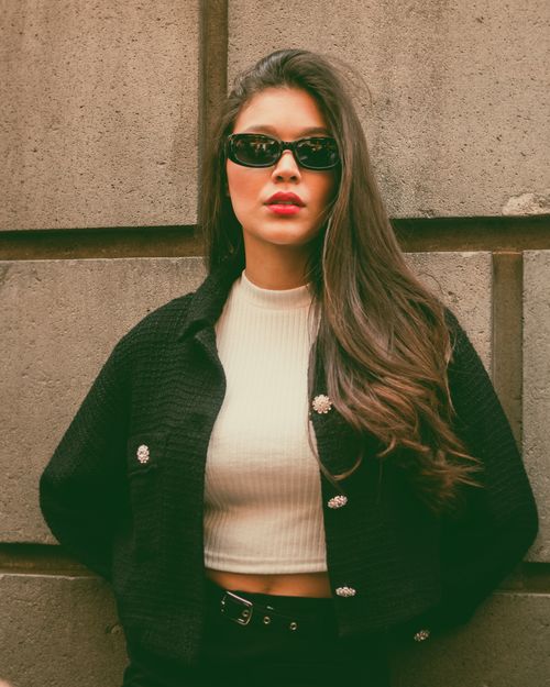 Free Stylish woman wearing a black jacket and sunglasses poses confidently against a wall. Stock Photo