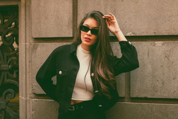 A woman in dark sunglasses and a tweed jacket posing against a stone wall — photo by Andrea Román on Pexels