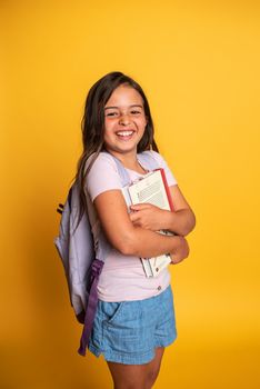 Cheerful girl with a backpack and books posing in a studio against a vibrant yellow background.