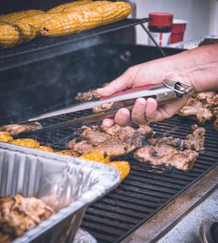 Close-up of chicken and corn being grilled, with a hand using tongs to barbecue.