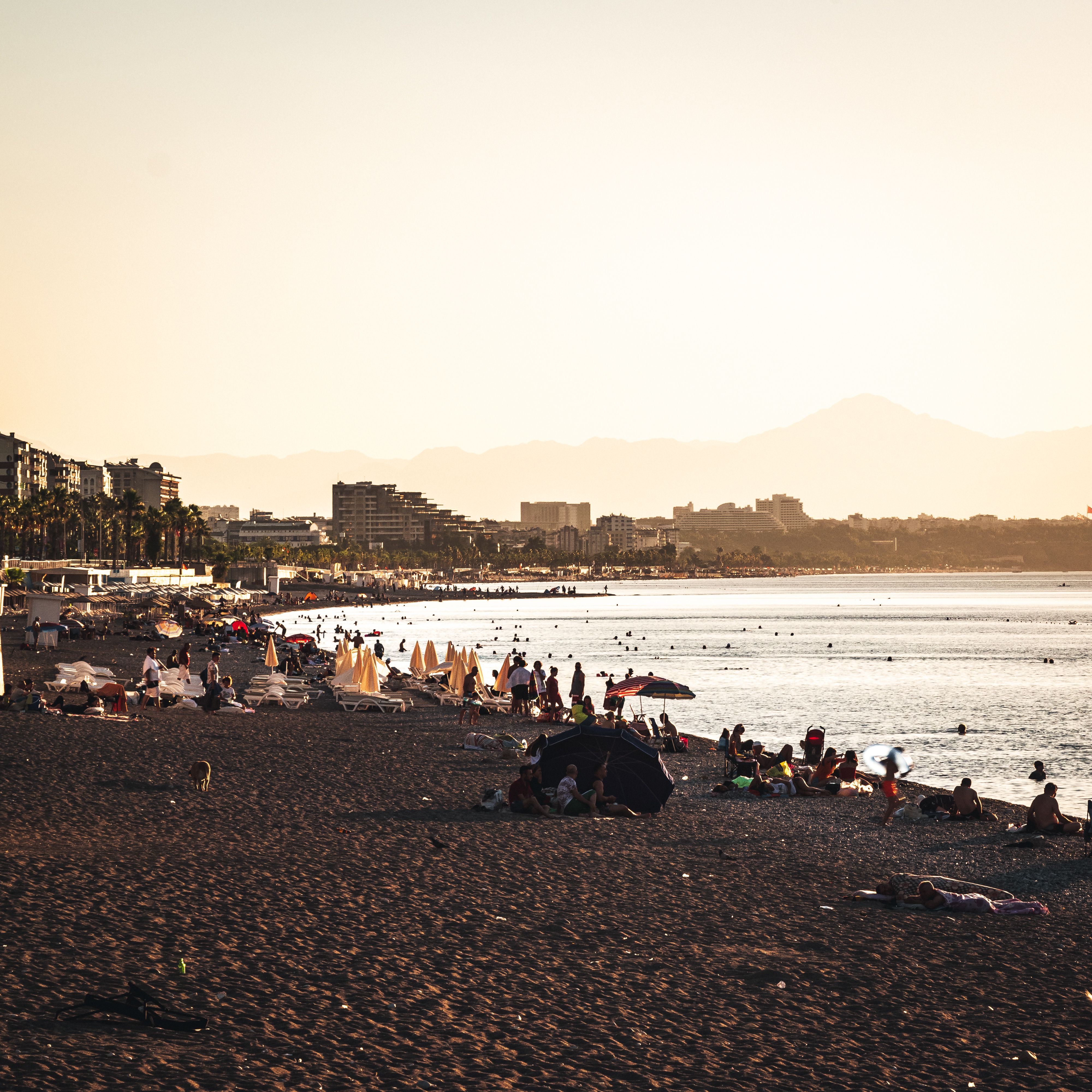Summer beach scene with people relaxing under sunset sky and mountains in the distance.