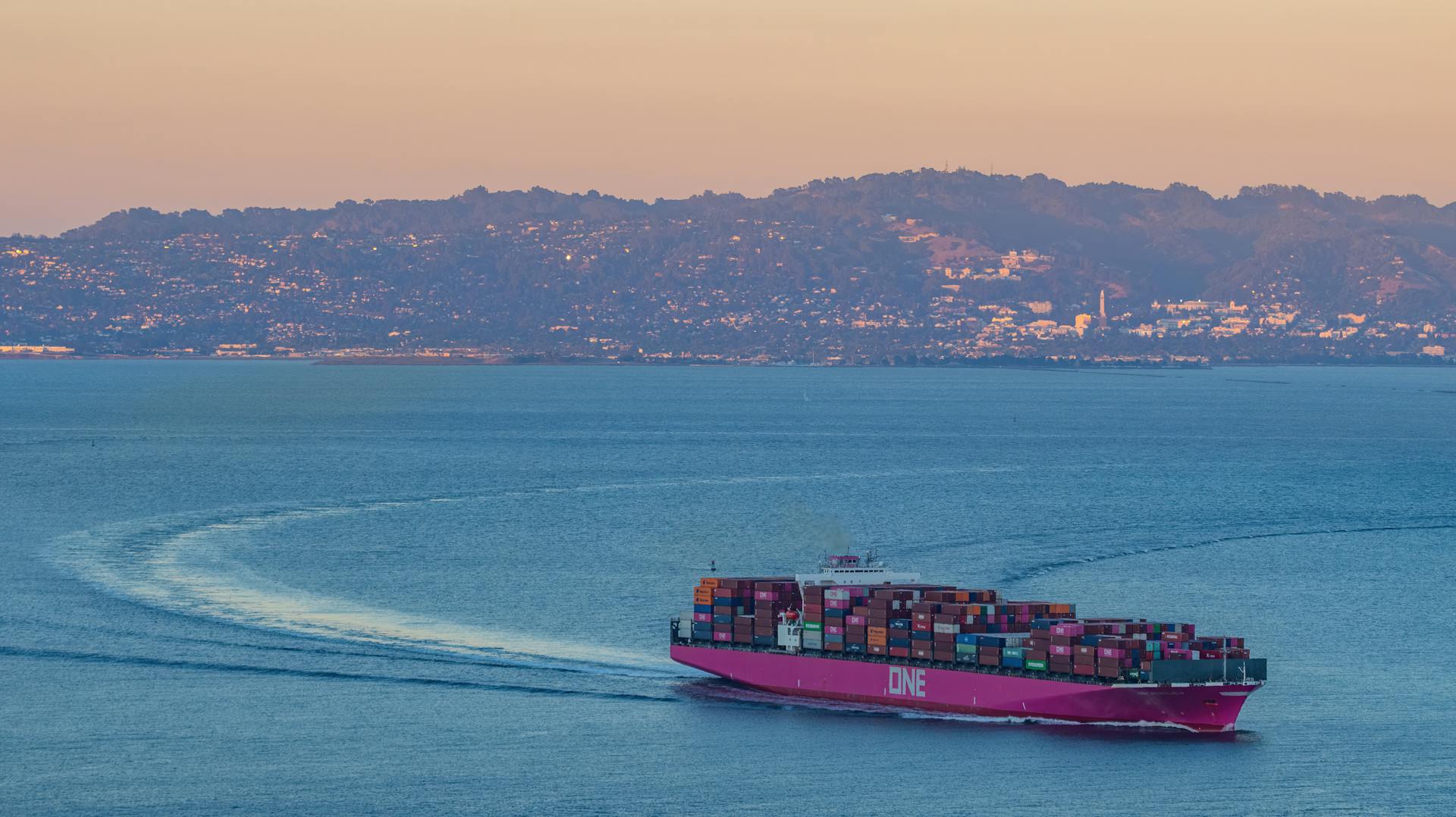 A cargo ship travels at sea with colorful containers against distant hills during sunset.