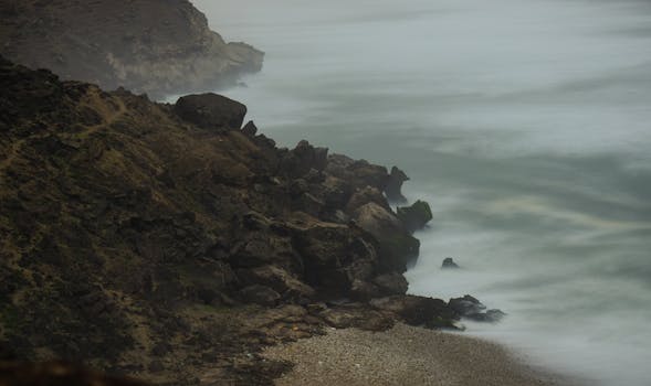 Misty ocean waves crash against a rocky shore under a foggy, gloomy sky.