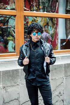 Trendy young man in black attire posing confidently against a colorful urban backdrop.