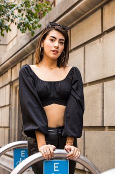 Fashionable young woman in a black crop top and hoop earrings posing confidently against an urban backdrop.