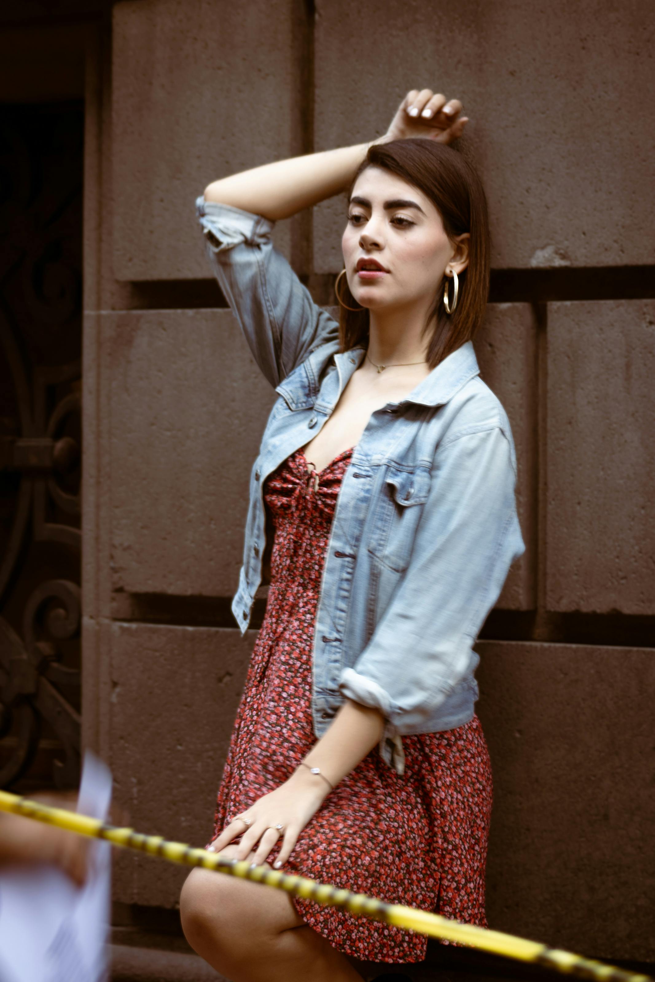 Stylish urban portrait of a woman in a denim jacket and floral dress posing against a wall.