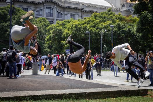Three men perform impressive parkour flips in a bustling urban park, showcasing athletic skills and artistry.