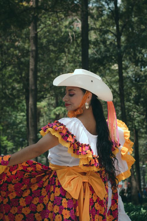 Free A woman in a vibrant floral dress and cowboy hat dances gracefully in a forest. Stock Photo