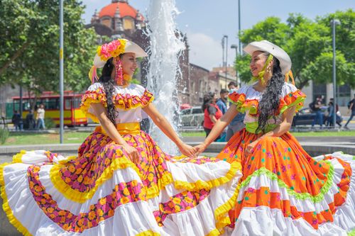 Free Two women in vibrant traditional Mexican dresses holding hands in front of a fountain. Stock Photo