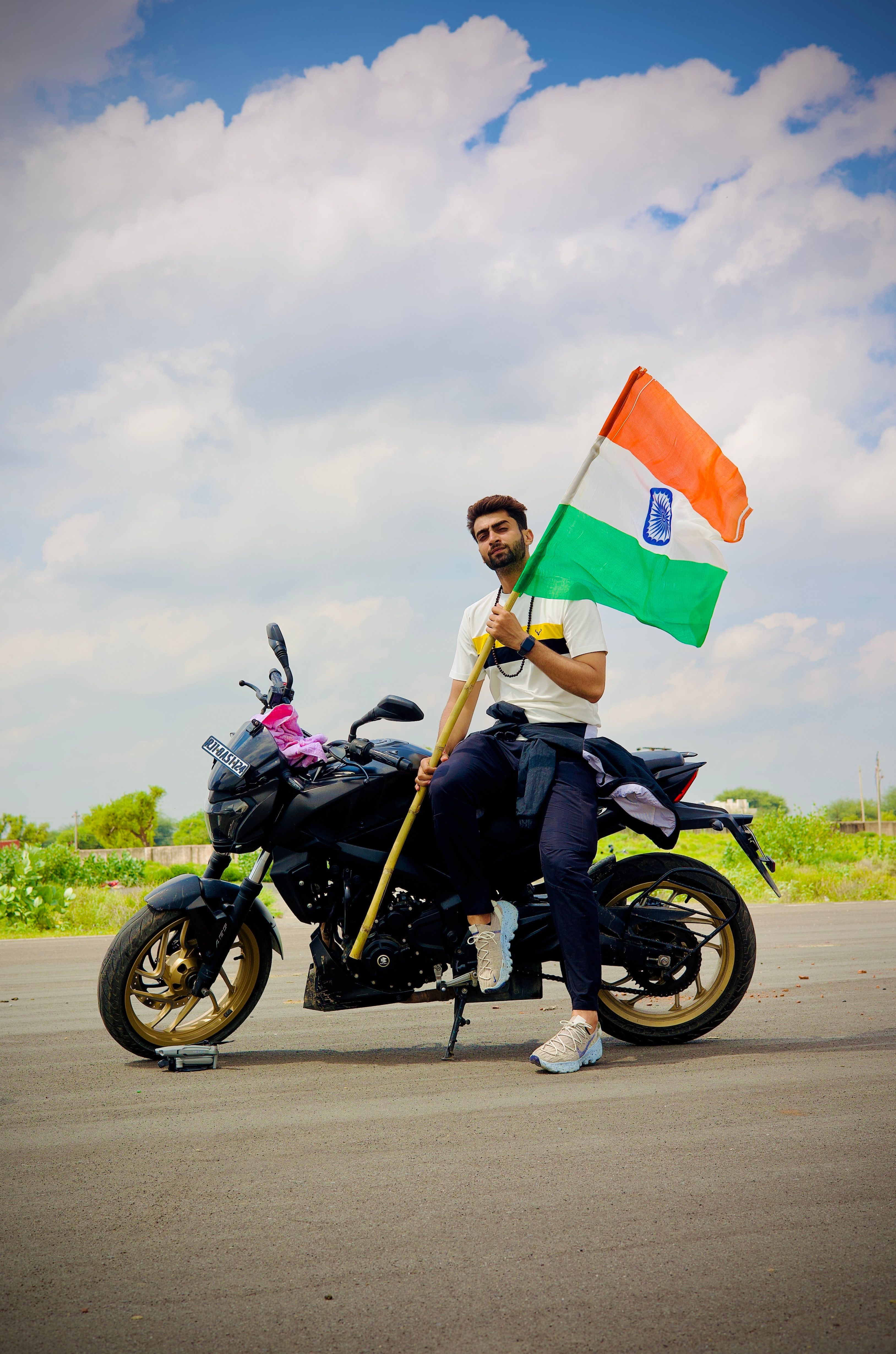 A man sitting on a motorcycle outdoors, proudly holding the Indian flag under a clear sky.