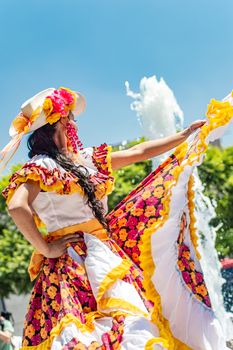 A woman in colorful traditional Mexican attire dances outdoors with a fountain in the background.