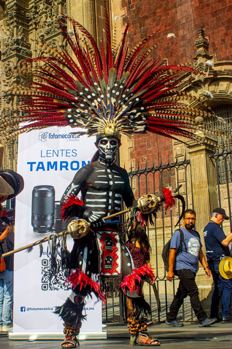 A Man With Halloween Make-up And Headdress Holding A Stick With Skulls