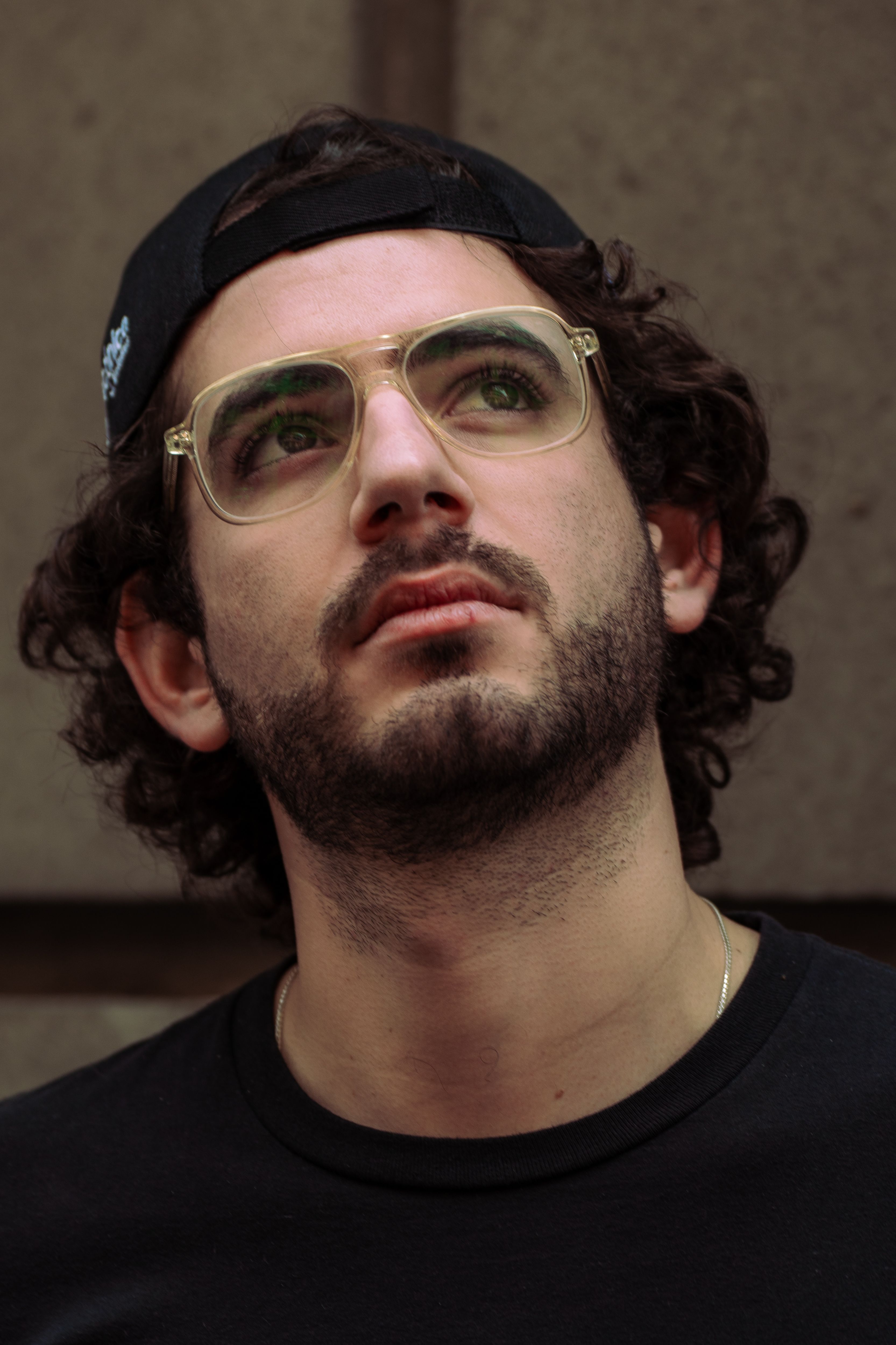 Close-up portrait of a bearded man in eyeglasses and cap, looking upwards outdoors.