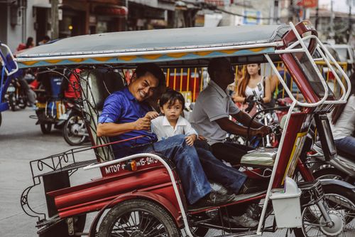 Free A man and child smiling while riding a colorful rickshaw in a vibrant urban setting. Stock Photo