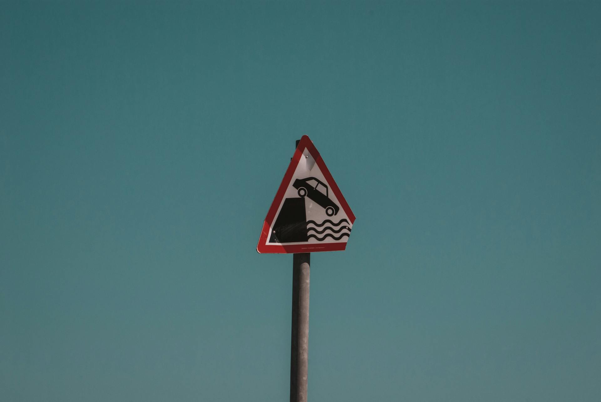 Triangular traffic warning sign showing a car falling into water, set against a clear blue sky in Dublin, Ireland.