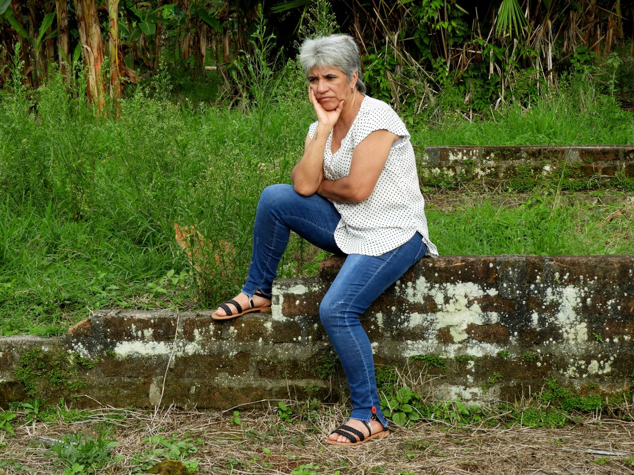 a mature woman in casual attire sits pensively on an old brick wall in
