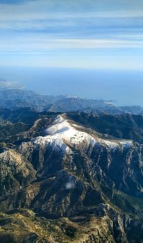 Breathtaking aerial view of a snow-capped mountain range with the sea in the distance under a clear blue sky.