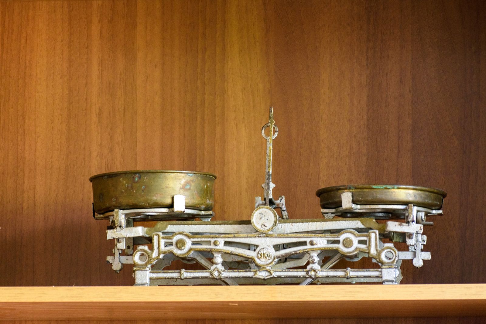 A vintage brass and iron balance scale with two brass pans at uneven heights on a wooden shelf