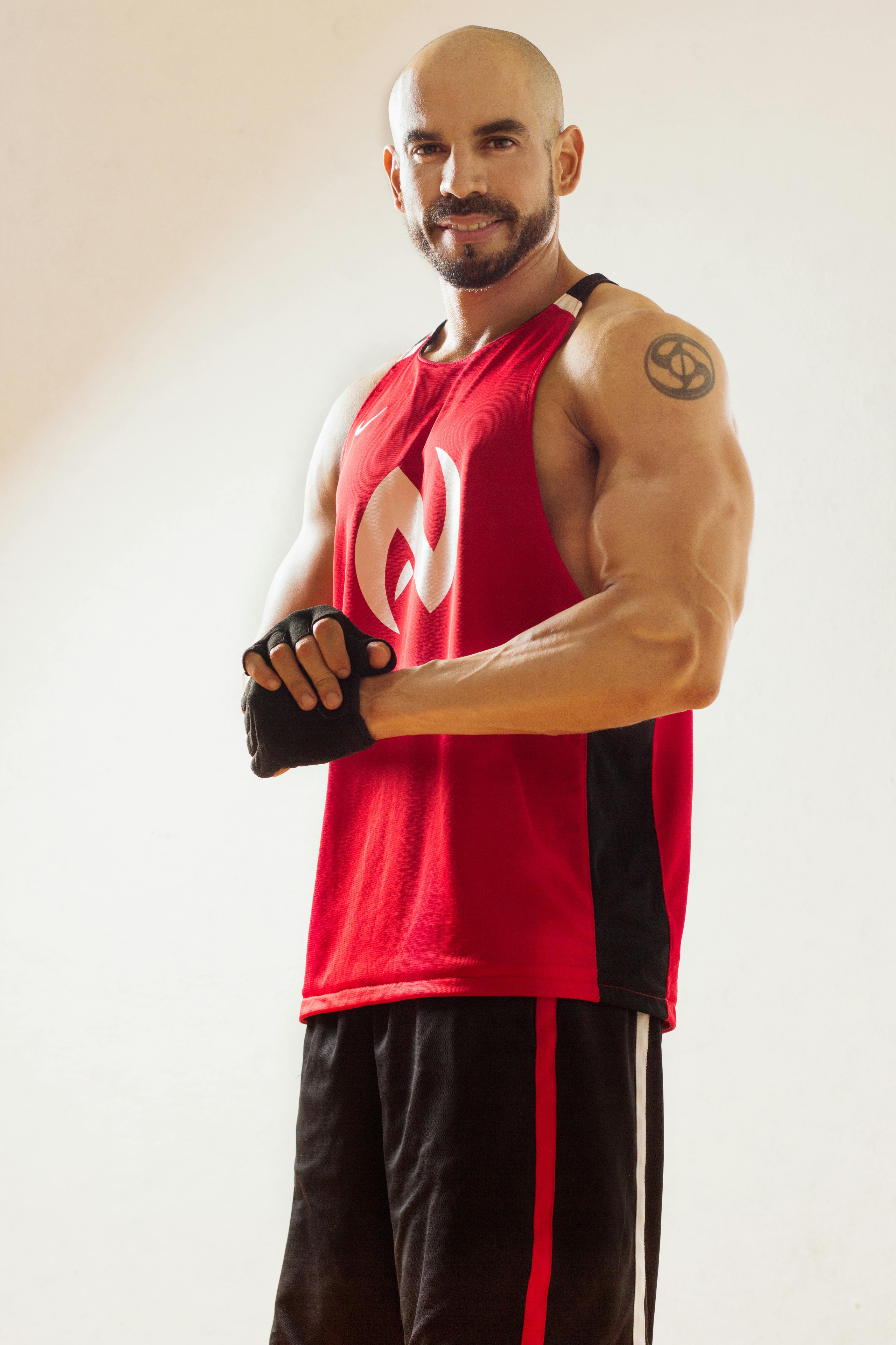 Athletic man in a red tank top, showcasing biceps and tattoos, smiling indoors.