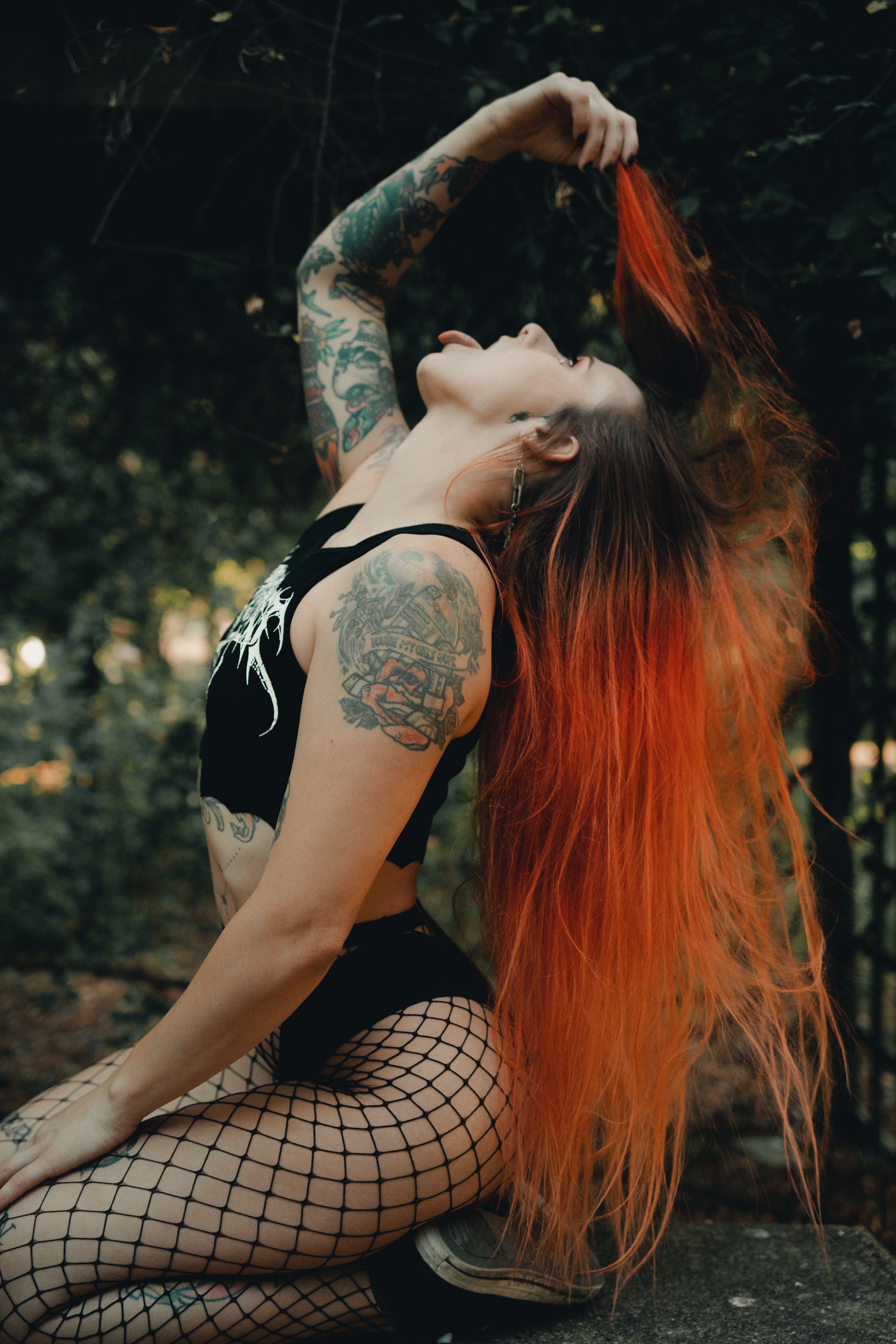 A woman with tattoos and dyed red hair kneels in nature, showcasing sensuality and style.