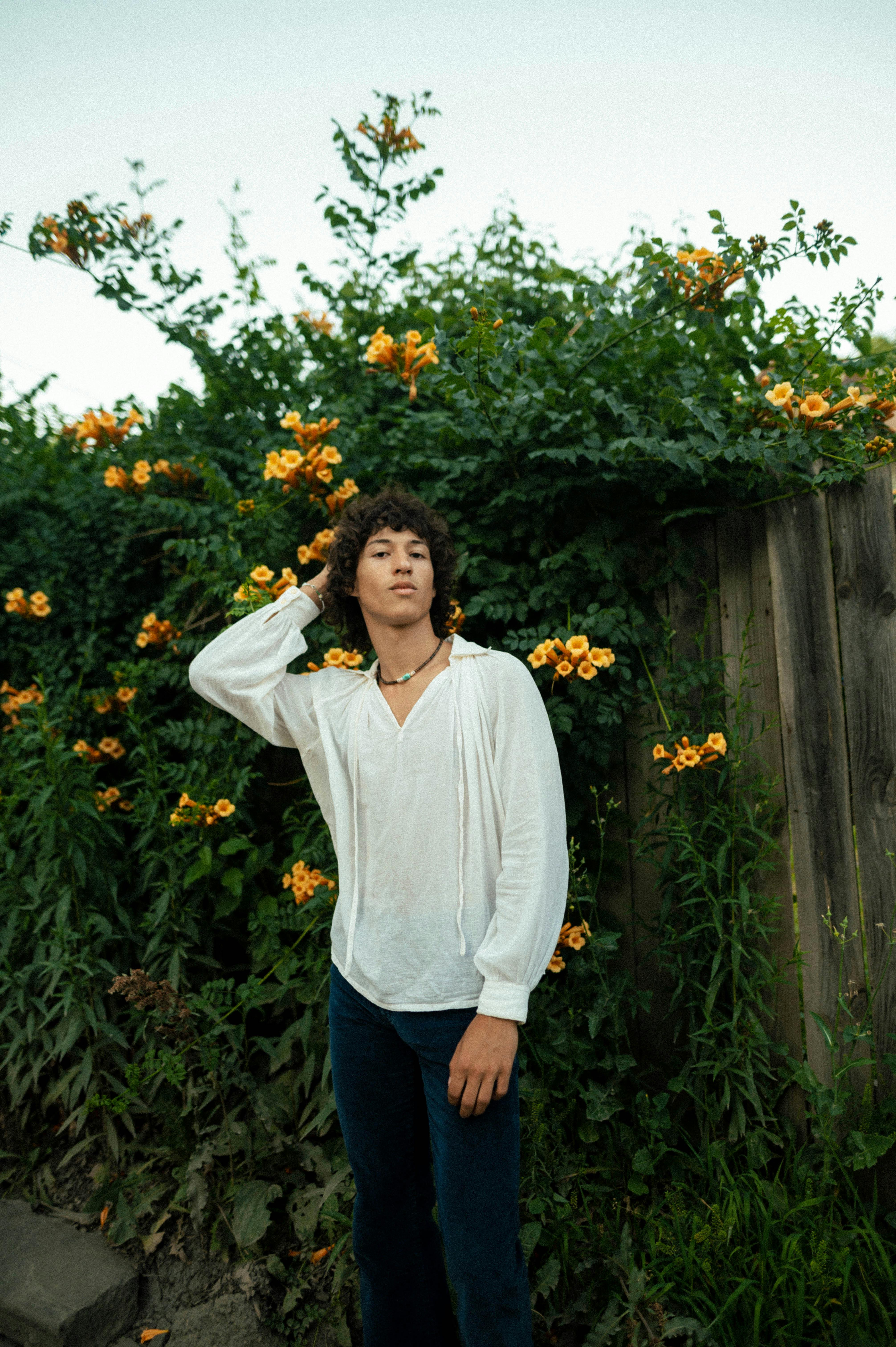 Stylish young adult in white shirt posing near a flower-covered fence outdoors.