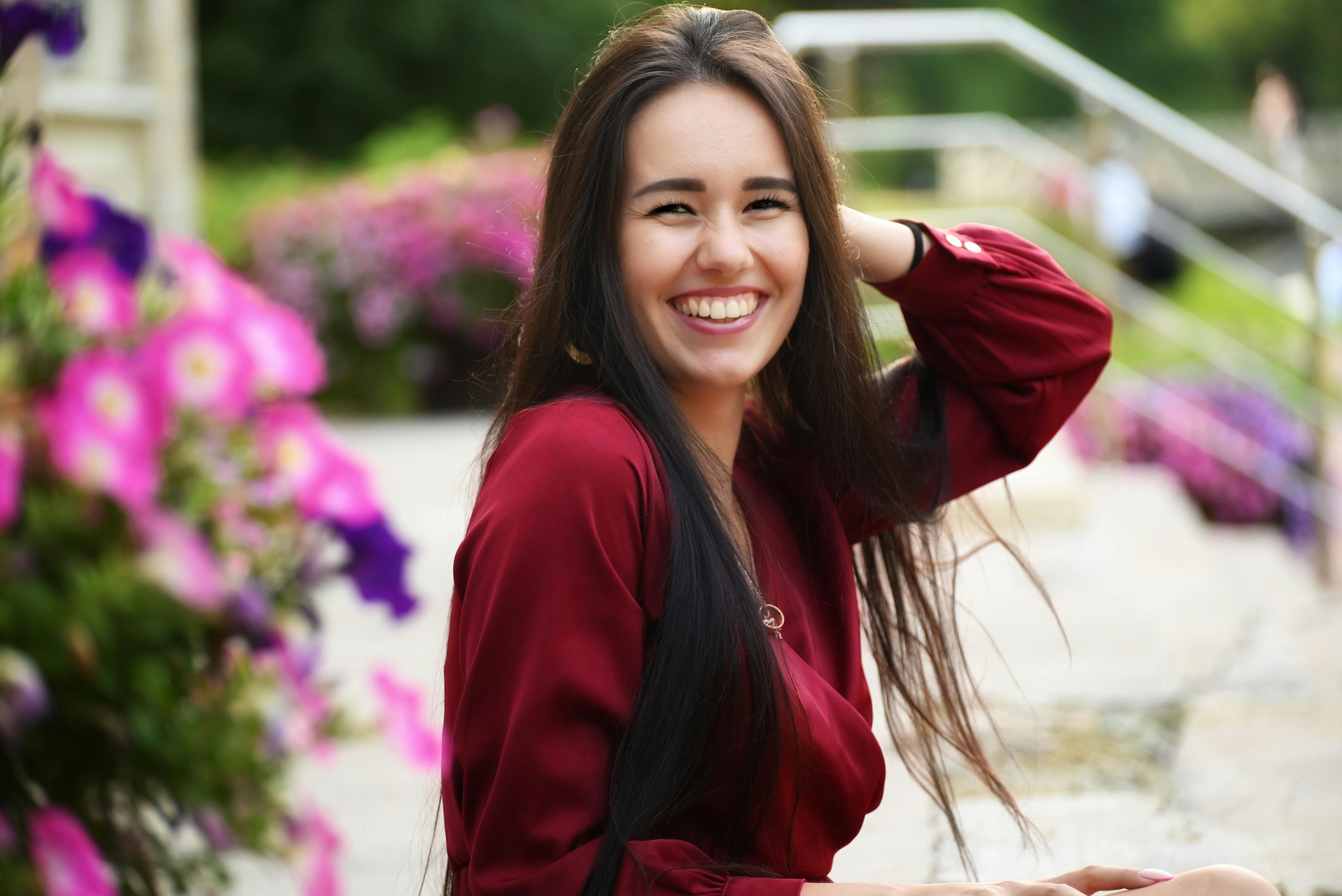 A cheerful young woman in a red dress posing outdoors with vibrant flowers.