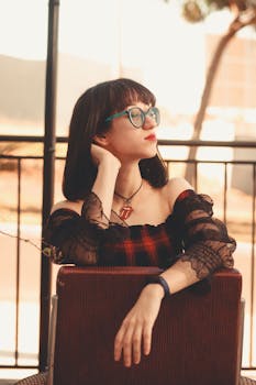 A fashionable young woman with eyeglasses poses leisurely outdoors in a chic outfit.