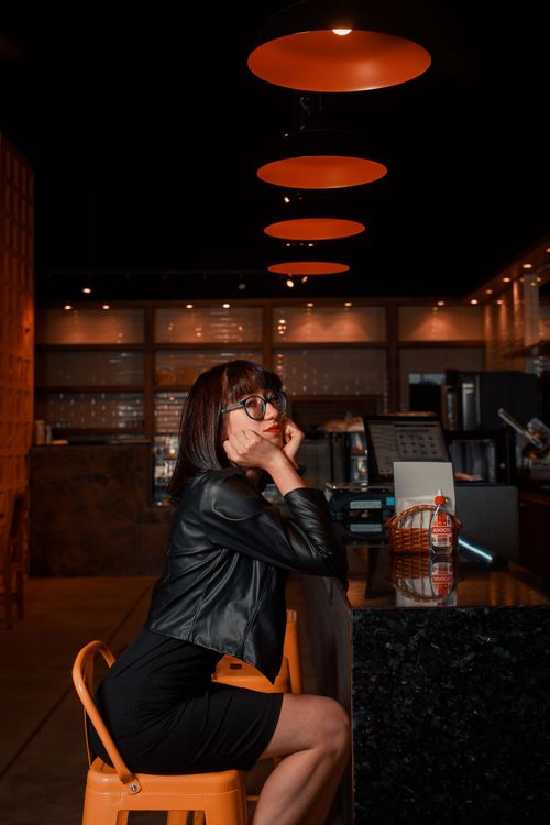Free An Asian woman in a black leather jacket sits thoughtfully at a bar counter under warm lighting. Stock Photo