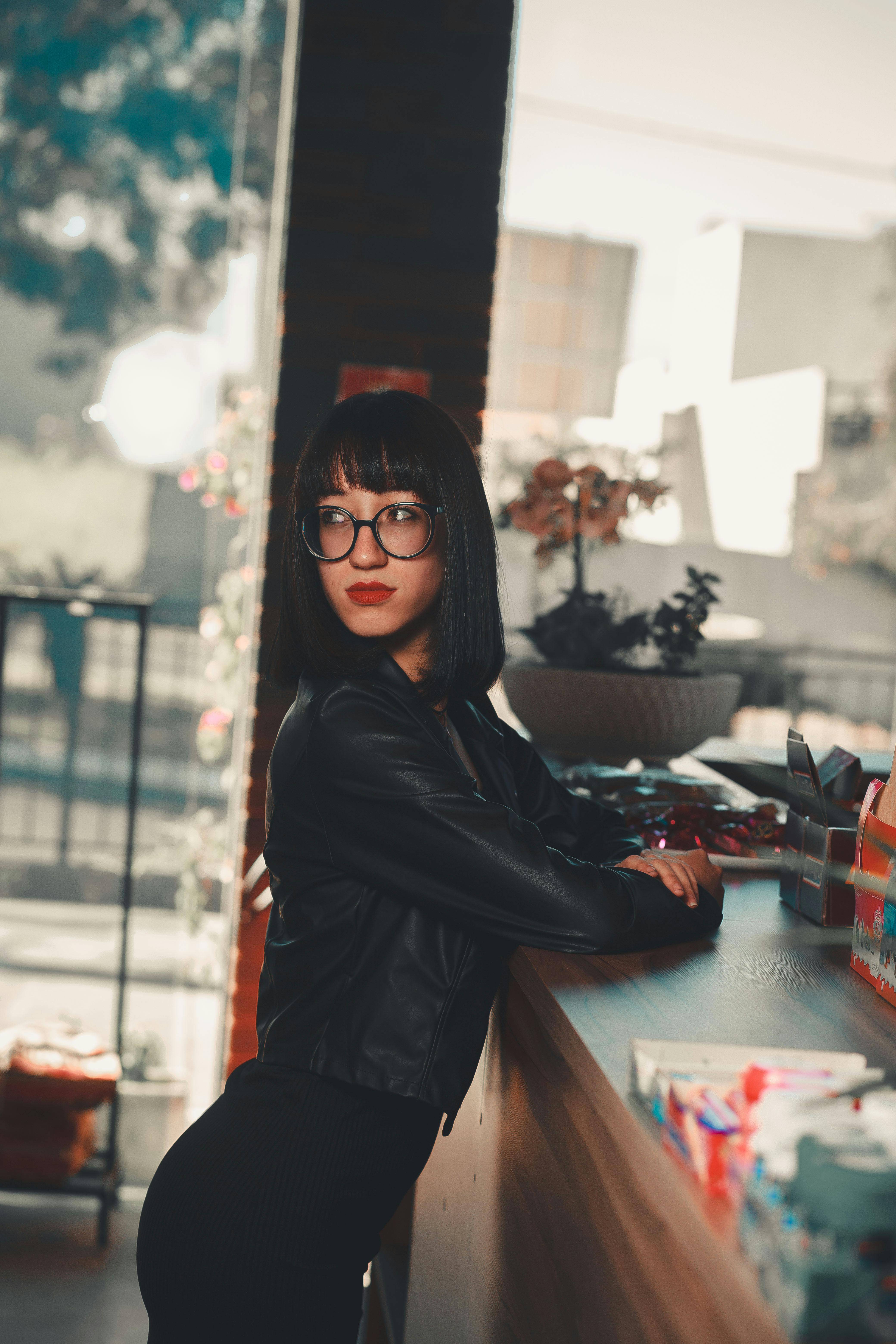 Fashionable woman in black leather jacket and glasses posing indoors.