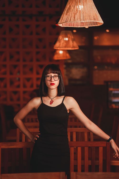 Free Portrait of a stylish young woman in a chic black dress and eyeglasses in a warmly lit indoor setting. Stock Photo