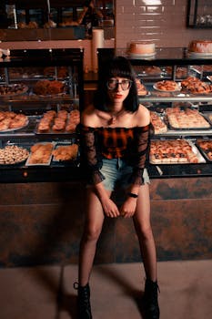 Stylish woman with eyeglasses posing in bakery surrounded by tempting pastries.