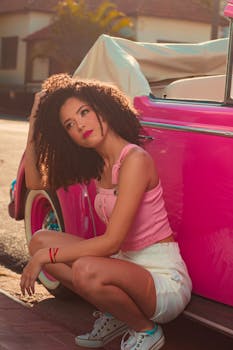 Fashionable woman crouching beside a vibrant pink vintage car on a sunny street.
