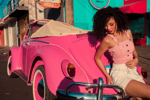 Stylish woman in retro outfit posing by a classic pink convertible car on a sunlit street.