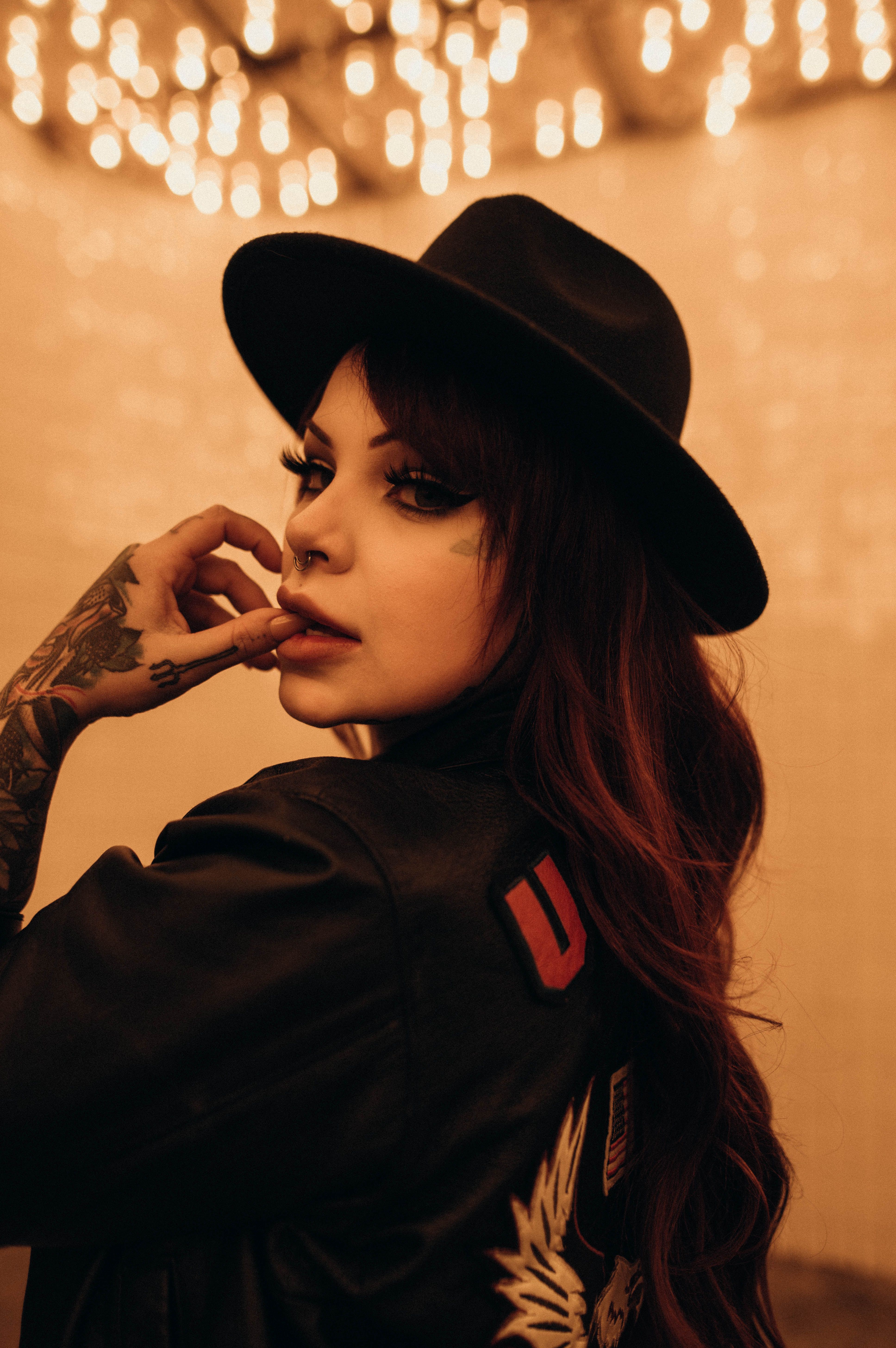 Tattooed woman in black hat posing indoors under warm lighting, showcasing modern fashion.