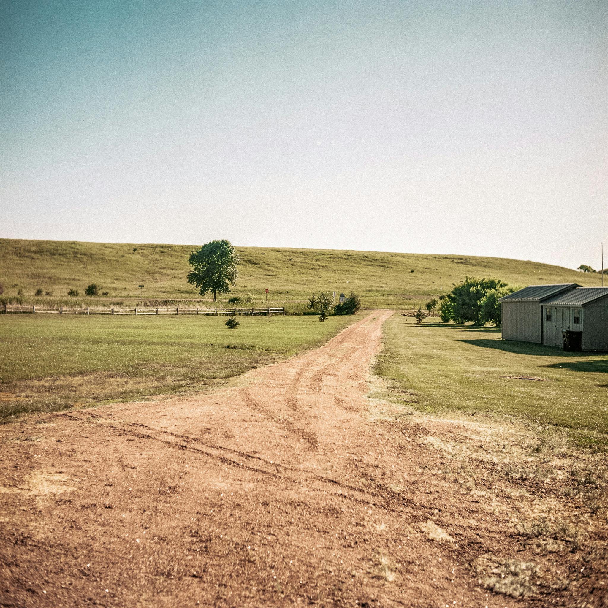 A quiet dirt road leads to a rural farm under a clear blue sky, capturing the essence of countryside life.