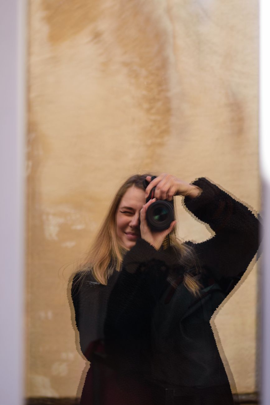 A woman holding a camera appears through a glass reflection, capturing her focused expression.