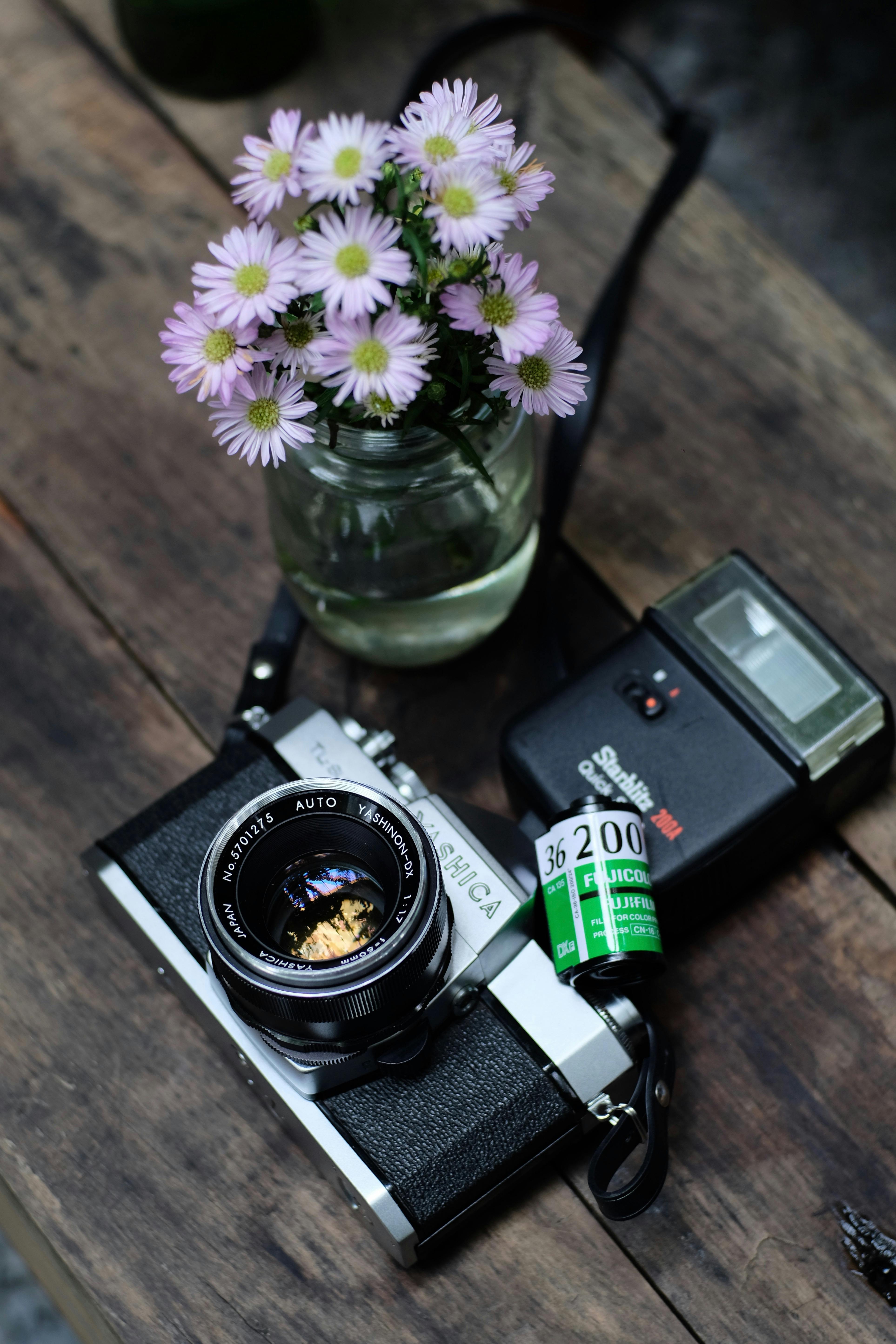 A vintage film camera with daisies in a jar on a rustic wooden table, featuring a retro photography theme.