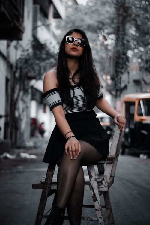 Free Fashionable woman sitting on chair in urban street, showcasing modern style with sunglasses and off-shoulder top. Stock Photo