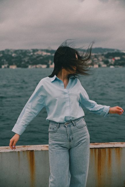 A woman standing by the ocean with her hair blowing in the wind, captured outdoors in a casual style.