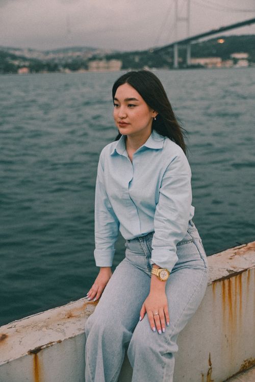 Free Woman sitting casually by the ocean with a bridge in the background, creating a serene mood. Stock Photo