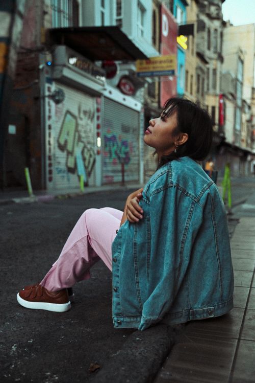 Free Young woman in denim jacket sits thoughtfully on a city street, surrounded by urban graffiti. Stock Photo