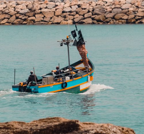 Free A vibrant fishing boat navigates through calm waters with fishermen on board, against a rocky shoreline. Stock Photo