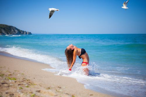 Free A woman in a bikini kneels on the beach with birds flying above, embracing the ocean's waves. Stock Photo