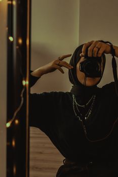 Young woman in black outfit and hijab taking a mirror selfie with accessories and soft lighting.